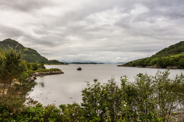 Stromeferry, Scotland - June 10, 2012: From Castle Strome ruins looking over Loch Carron towards ocean. Green hills on shores. Motor boat anchored in lake at distance. Rain-heavy cloudscape.