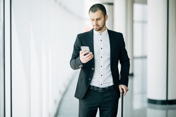 Elegant businessman checking e-mail on mobile phone while walking with suitcase inside airport terminal. Experienced male employer using cell telephone before work travel