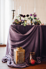 Festive table decorated by violet cloth, flowers and greenery, candles on candleholders with golden lantern and candles near them. Wedding decor