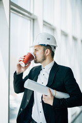Engineer drink coffee with blueprint in hands before inspection of construction against panoramic windows