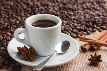 a cup of espresso on a saucer with a spoon and aniseed cinnamon sprigs and roasted coffee beans