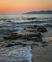 A long exposure of the sea as it breaks over the rocks and beach at golden hour, with the sun setting behind the mountains.