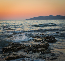 Fototapeta premium A long exposure of the sea as it breaks over the rocks and beach at golden hour, with the sun setting behind the mountains.