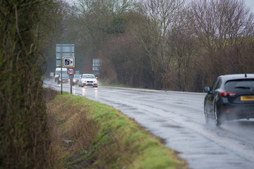 cars driving along water logged road with spray and rain and camera and 50 mph speed warning sign