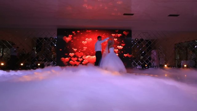 The beautiful happy couple of newlyweds are dancing their first dance at the background of the screen display with hearts and are shrouded by the white fume. Restaurant location.
