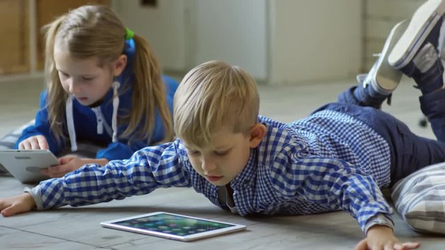 Adorable blonde boy and girl lying on the floor in kindergarten and surfing the Net on digital tablet