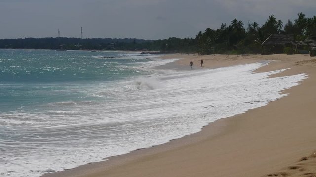 People walk barefoot along the ocean and a wide sandy beach in the tropics