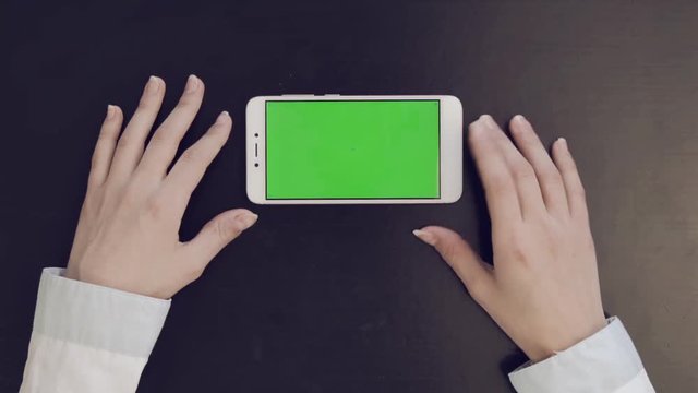 Green screen on smartphone. Businesswoman in office at her desk and works with phone. View from top. Looks and scrolls her finger from bottom to top of mobile app menu. Phone horizontally.