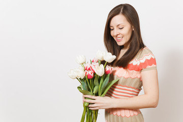 Beautiful young brunette woman in light patterned dress holding bouquet of white and pink tulips in hands, rejoices and looks at flowers in studio on white background. Concept of holiday, good mood.