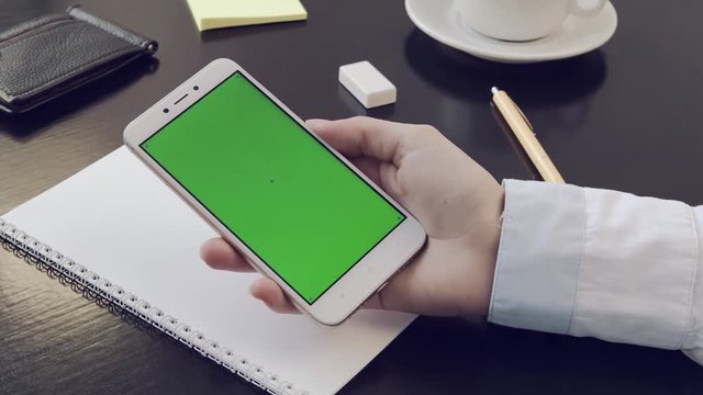 Green screen on smartphone. The businesswomen 's hand is holding the phone at the desk in the office. Scroll horizontally on the phone screen. Jacket, women 's hand and phone.