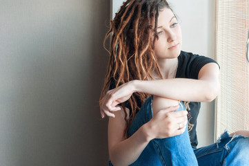 Portrait of a young girl in dreadlocks and torn jeans sitting on the window of the house