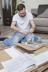 Mężczyzna klęczy na dywanie nad kartonem z elementami mebli do samodzielnego montażu i rozcina nożyczkami torebkę foliową z wkrętami i śrubami do montażu.