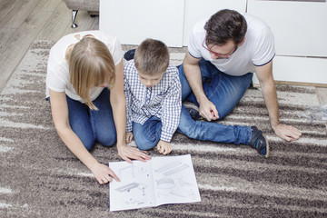 Rodzina klęczy na beżowym dywanie. Przed nimi leży instrukcja montażu mebli, którą czytają z wielką uwagą. - obrazy, fototapety, plakaty