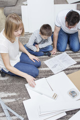 Tata z synem wpatrują się w instrukcję montażu mebli klęcząc na dywanie. Mama wybiera jeden...