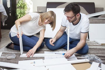 Praca zespołowa. Mąż i żona skręcają razem meble w salonie dopasowując drewniane  elementy zestawu meblowego. - obrazy, fototapety, plakaty