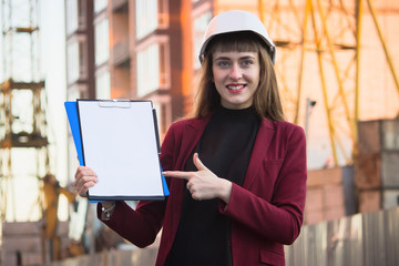 Woman builder holding blueprints, clipboard. Smiling architect girl in helmet at building background