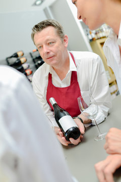 Man in apron showing bottle of wine to couple