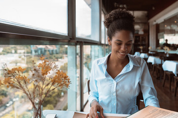 Dainty Afro American girl in a teal shirt is sitting inside of a luxury restaurant near the window and choosing food to order from the menu; black young female in cafe is rifling the pages of a menu - Powered by Adobe