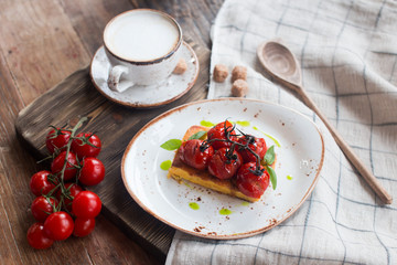 Fototapeta na wymiar Traditional Italian vegetarian focaccia with fried cherry tomatoes, cappuccino cup with pieces of cane sugar, table covered with a tablecloth with a wooden spoon close up