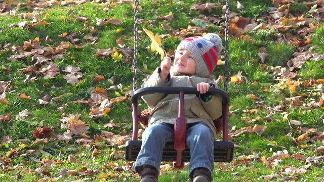 Adorable child with a yellow leaf smiling on swing, beutiful autumn