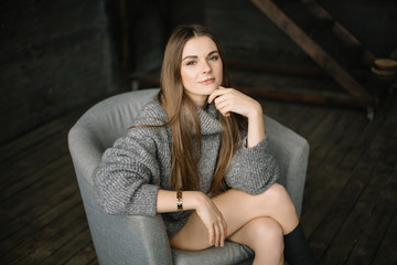Obraz premium Dark haired sad woman in long grey knitted cozy sweater sitting in a chair at home near big window. Happy and Healthy girl with long beautiful legs with soft skin in studio. Daylight, serious teen.