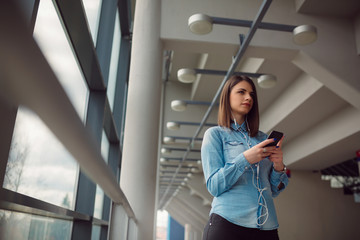 Confident beautiful business woman looking aside and using smartphone in light office hall. Girl listening to the music on airport.