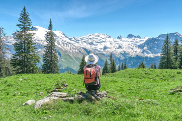 Swiss Alps. A woman in a white hat is sitting on a green meadow, admiring the mountain scenery....