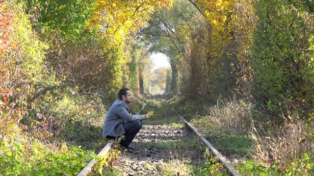 Single man sitting on railway playing with leaf, trees tunnel, autumn sunny day