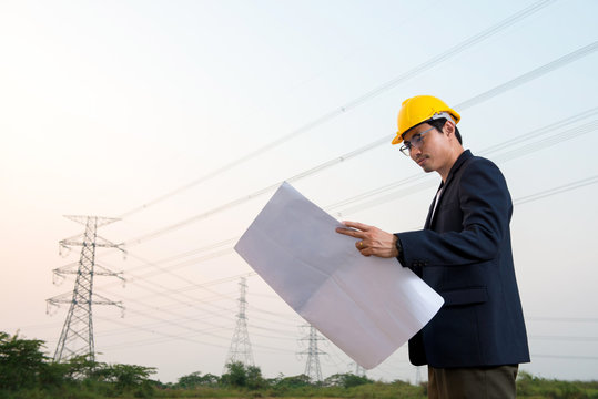 An engineer wear yellow hard hat and suite hold blueprint in hand standing on field looking at blueprint