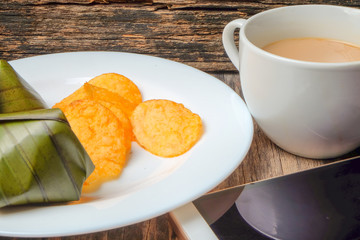 A cup of coffee, crispy potato chips and koci the malay glutinous rice ball served in banana leaf on white plate over wooden table