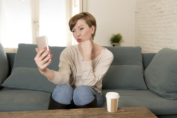 young attractive and happy red hair woman sitting at home sofa couch drinking coffee taking selfie picture with mobile phone camera