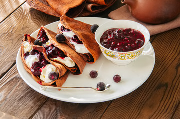 Chocolate crepes (blinis) with sour cream and black currant sauce on a white plate. Wooden...
