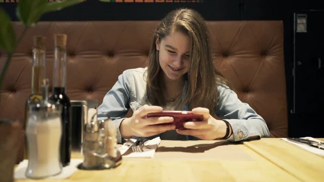 Happy teenage girl playing game on smartphone sitting by table in cafe
