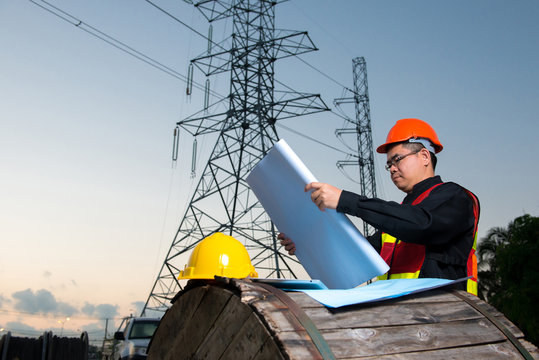 Electrical engineer working. Electrician holding blueprint at high voltage power pylon against blue sky