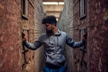 Obraz premium Portrait of focused motivated afro-american young handsome sportive man with earphones standing inside of the abandoned place in the middle of two walls and thinking while looking aside.