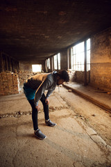 Fototapeta premium Portrait of active afro-american young attractive athletic man with earphones crouching and resting after workout inside of the abandoned place.
