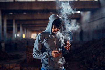 Portrait of joyful afro-american young handsome man relaxing inside of the abandoned hangar place with earphones and cigarette while choosing music on a mobile.
