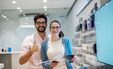Portrait view of pretty excited charming smiling young student girl with eyeglasses reading specification while her boyfriend standing next to her and showing thumb up and looking at the camera.