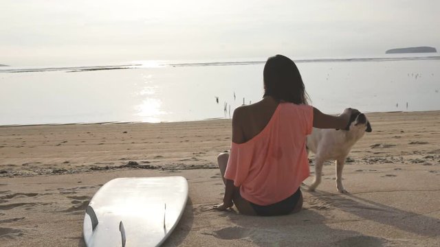 young brunette girl or woman in shape resting on sunny coast, caressing white dog, white surfboard is on the ground (sand). sunny day, ocean or sea, awesome nature