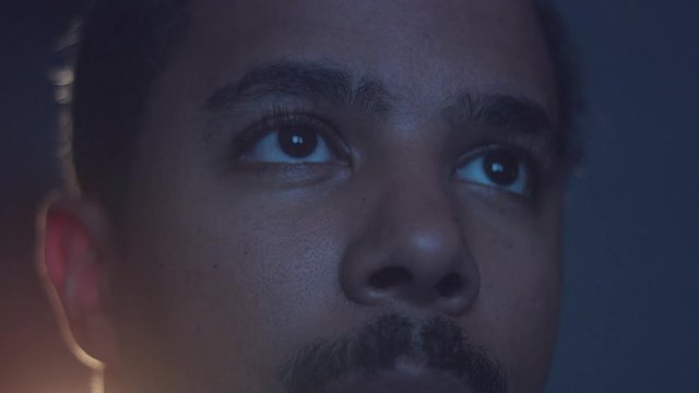 upset boring mixed race man watching tv in darkness with different color light from tv on face. Eyes closeup