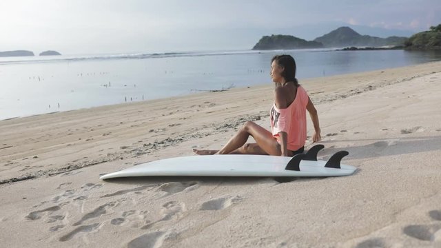 smiling happy sporty girl or young woman or female surfer enjoying her time on the beach or coast, looking away. calm sea or ocean, hills or mountains, sandy beach or coastline.