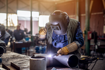 Profesional welder in protective uniform and mask welding metal pipe on the industrial table with other workers behind in the industrial workshop.
