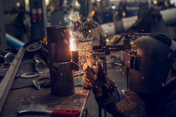 Welder in protective uniform and mask welding metal pipe on the industrial table with other tools while sparks flying.