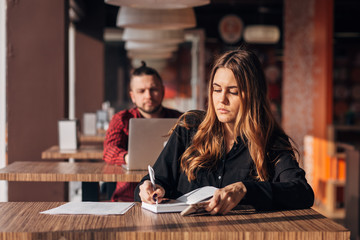 business young girl writing note in notebook in cafe, man with laptop on background