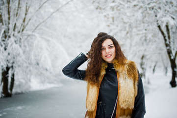 Elegance curly girl in fur coat at snowy forest park at winter.
