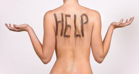 A young girl poses nude in front of the camera. She has  her back turned away from the camera. She has the word  help  painted in black on her back.