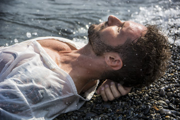 Handsome muscular man on the beach lying on gravel, wearing wet white shirt
