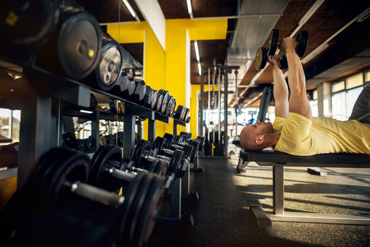 Side view of strong motivated and focused muscular bald bodybuilder man doing chest exercise while lying on the bench with dumbbells and row of dumbbells behind him in the modern sunny gym.
