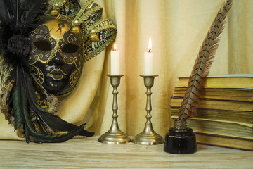 Literature concept, candle in a candlestick near a Venetian mask and an inkwell with a pen, books on a light background