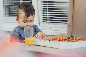 the little boy eats a huge harmful pizza himself in the kitchen and drinks juice, very fat and...